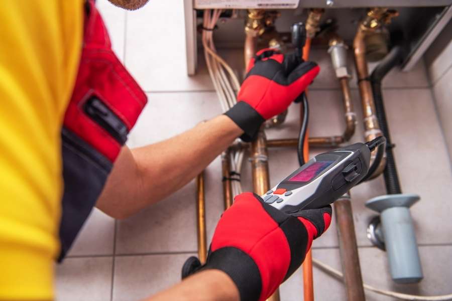 Un technicien portant des gants rouges et noirs utilise un appareil de mesure pour inspecter des tuyaux et des raccordements de plomberie.